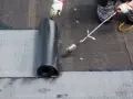 Pracownik w rękawicach wykonuje układanie papy na zimno, podgrzewając ją palnikiem.