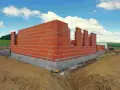 Budowa domu w toku: czerwone ściany z pustaków, fundamenty z betonu i bloczków. W tle zielone pole i błękitne niebo z chmurami.