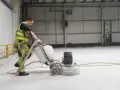 Pracownik w kamizelce odblaskowej wykonuje szlifowanie betonu za pomocą maszyny.