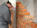 Murarz w kasku i rękawicach sprawdza pion muru z cegły, dbając o prawidłowe wymiary cegły i równość ścian.