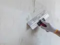 Ręka w rękawicy wykonuje szpachlowanie ścian, wygładzając nierówności.