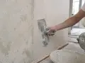 Ręka z kielnią nakłada gładź na chropowatą ścianę, przygotowując ją do malowania.