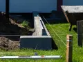 Budowa fundamentu pod ogrodzenie. Betonowe bloczki tworzą solidną podstawę, a sznur wyznacza linię. W tle taczka i zielona trawa.