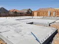 Fundament domu z chudego betonu, z widocznymi zbrojeniami i instalacjami. W tle powstaje drewniana konstrukcja domu.