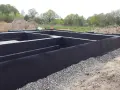 Fundament domu w trakcie budowy, zabezpieczony czarną membraną i otoczony żwirem.