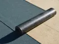 Rolka papy z połyskującą folią aluminiową leży na ciemnozielonej powierzchni.