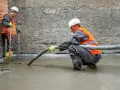 Dwóch budowlańców w kamizelkach odblaskowych wylewa pianobeton. Jeden kieruje wąż, drugi kontroluje poziom.