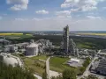 Zakłady Lafarge z lotu ptaka, z dominującą wieżą pieca cementowego i silosami, otoczone zielenią i polami.