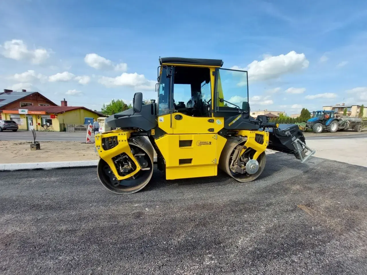 Żółty walec drogowy ugniata świeży asfalt. W tle widać budynki i niebieski traktor.