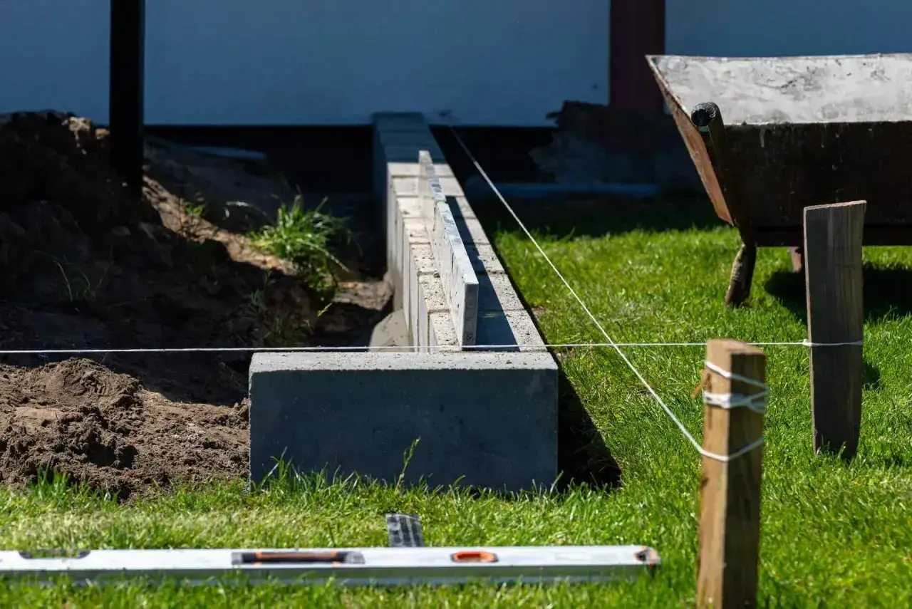 Budowa fundamentu pod ogrodzenie. Betonowe bloczki tworzą solidną podstawę, a sznur wyznacza linię. W tle taczka i zielona trawa.