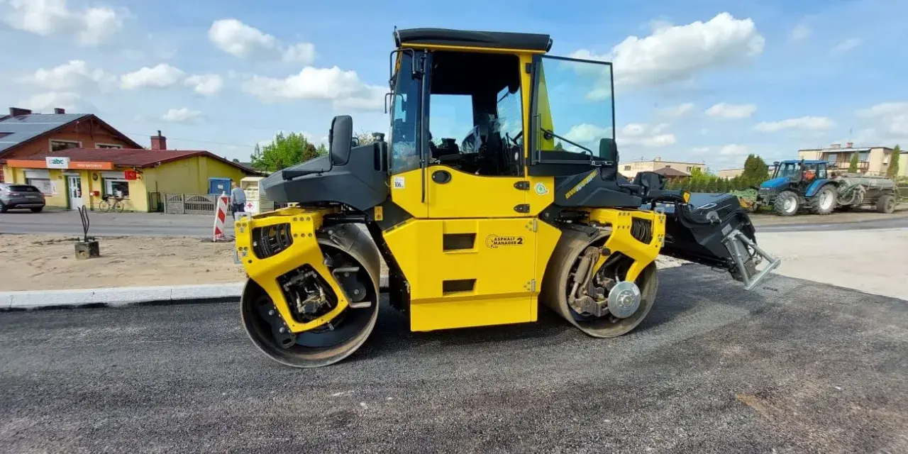 Żółty walec drogowy ugniata świeży asfalt. W tle widać budynki i niebieski traktor.