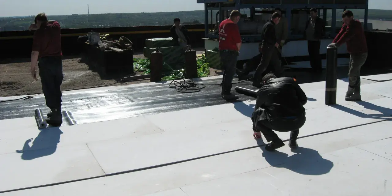 Mężczyźni pracują na dachu, układając materiał izolacyjny. W tle widać klimatyzator mgo.