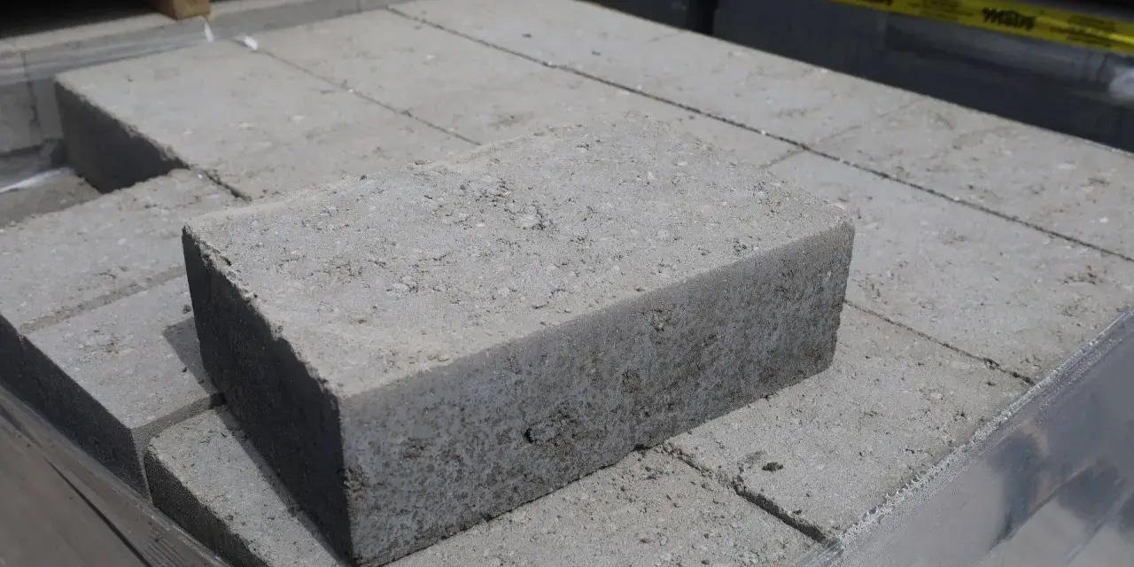 Stos szarych bloczków betonowych, każdy z widocznymi nierównościami i kruszywem. Wymiary bloczków betonowych są standardowe dla budowy.