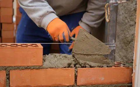 Budowa murowanego prostego grilla z cegły. Ręce w pomarańczowych rękawiczkach nakładają zaprawę kielnią.