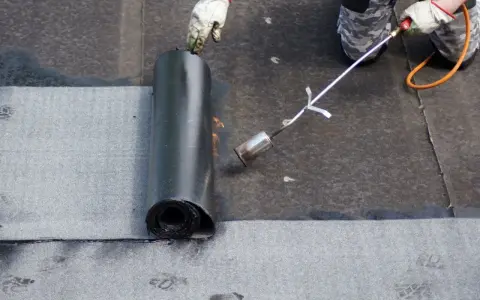Pracownik w rękawicach wykonuje układanie papy na zimno, podgrzewając ją palnikiem.