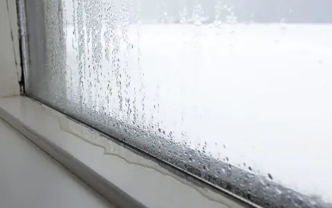 Intensywne skraplanie na oknie, krople wody spływają po szybie i tworzą kałużę na parapecie.