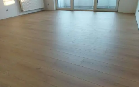 Jasne panele ułożone wzdłuż pomieszczenia, tworząc wrażenie większej przestrzeni. W tle biały grzejnik i okno balkonowe.