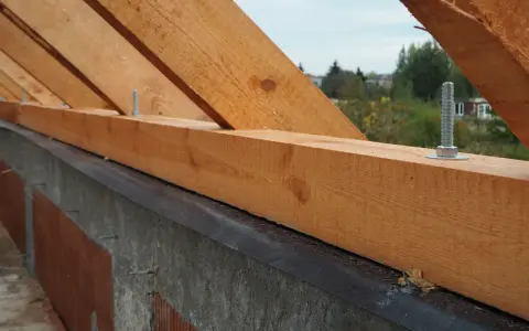 Drewniana murłata z widocznymi wymiarami, przykręcona do betonowej ściany. W tle drzewa i domy.