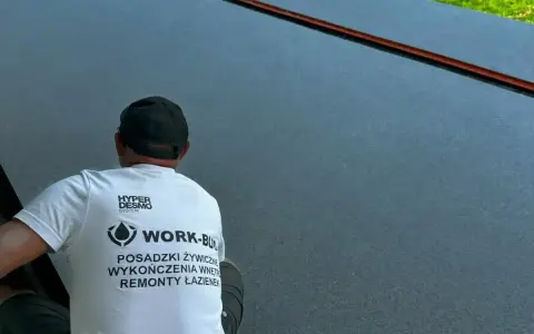 Pracownik w koszulce z napisem "Posadzki żywiczne" wykonuje prace na ciemnej, gładkiej powierzchni.