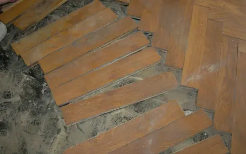 Drewniana podłoga w jodełkę, częściowo rozebrana. Widać pojedyncze deseczki, gotowe do ułożenia lub demontażu. Subit, klej do parkietu, wciąż widoczny na niektórych elementach.