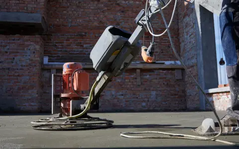 Robot do zacierania betonu pracuje na budowie. Robotnik w niebieskich spodniach kieruje maszyną, która wygładza świeżą posadzkę.