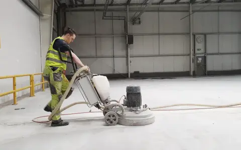 Pracownik w kamizelce odblaskowej wykonuje szlifowanie betonu za pomocą maszyny.
