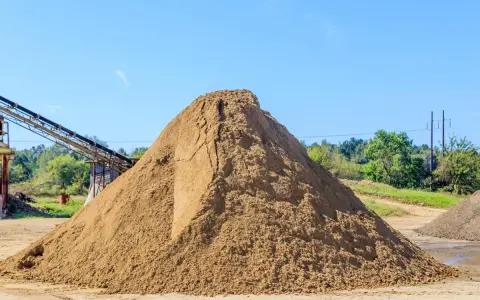 Duża hałda piasku na placu budowy, z taśmociągiem w tle.