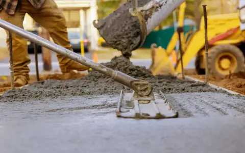 Pracownik rozprowadza świeży beton c12/15 za pomocą łaty. W tle widać koparkę i plac budowy.