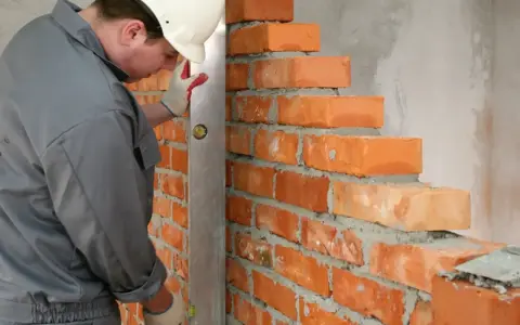 Murarz w kasku i rękawicach sprawdza pion muru z cegły, dbając o prawidłowe wymiary cegły i równość ścian.