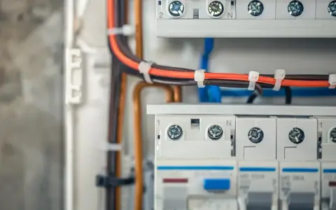 Szczegółowy widok instalacji elektrycznej w domu: przewody w kolorach czarnym i pomarańczowym, zabezpieczone opaskami, prowadzą do białej skrzynki z bezpiecznikami.