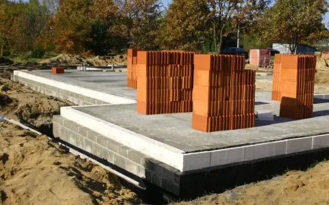 Stosy cegieł na budowie domu. Fundament z bloczków betonowych i płyta betonowa. W tle drzewa jesienią.