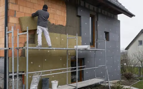 Pracownik montuje izolację na elewacji domu. Widać ceglaną ścianę, wełnę mineralną i szare płyty izolacyjne.