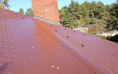 Czerwony dach pokryty gontem bitumicznym z widocznym kominem i drzewami w tle.