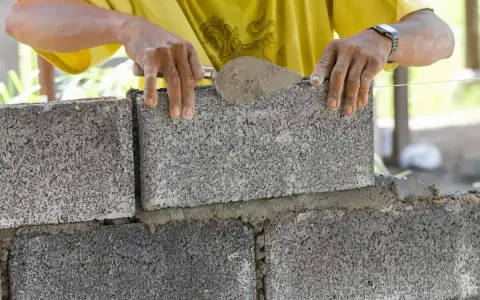 Budowniczy układa bloczki betonowe, tworząc ścianę. Widać, ile bloczków betonowych na m2 potrzeba do budowy.