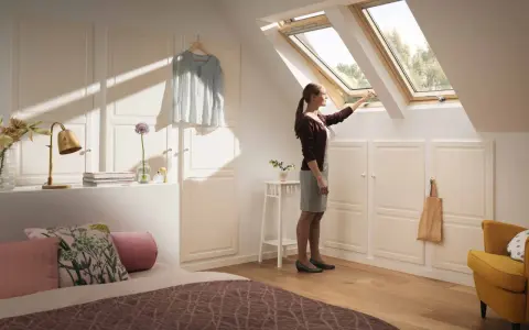Kobieta otwiera okno dachowe w pokoju z zabudową na ściankach kolankowych.