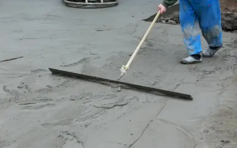 Pracownik w niebieskich ogrodniczkach wygładza świeży beton długą pacą. Trwa wygrzewanie posadzki.