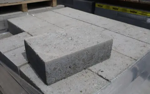 Stos szarych bloczków betonowych, każdy z widocznymi nierównościami i kruszywem. Wymiary bloczków betonowych są standardowe dla budowy.