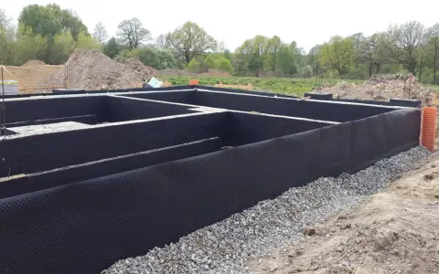 Fundament domu w trakcie budowy, zabezpieczony czarną membraną i otoczony żwirem.