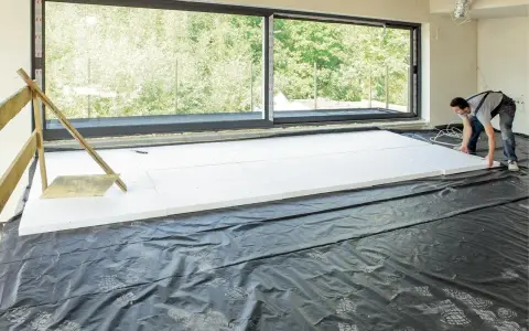 Pracownik układa płyty styropianowe do ocieplenie stropu betonowego. Duże okno wychodzi na zieleń.