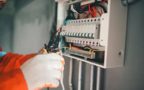 Elektryk w białych rękawiczkach montuje zabezpieczenie przedlicznikowe w skrzynce z bezpiecznikami.