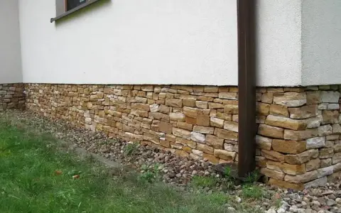 Eleganckie cokoliki z kamienia ozdabiają dom, tworząc naturalną, kamienną opaskę wokół białej ściany.