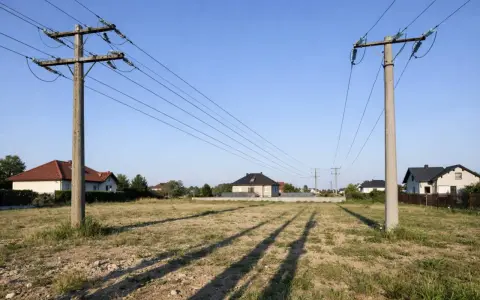 Słup energetyczny na działce, z przewodami biegnącymi nad polami i domami.