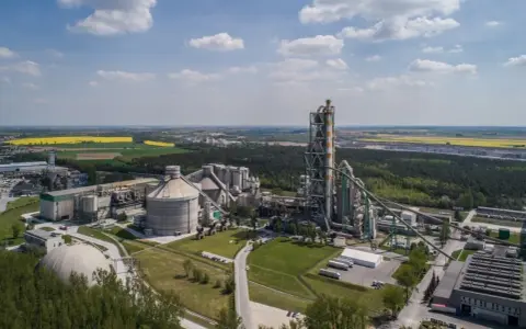 Zakłady Lafarge z lotu ptaka, z dominującą wieżą pieca cementowego i silosami, otoczone zielenią i polami.
