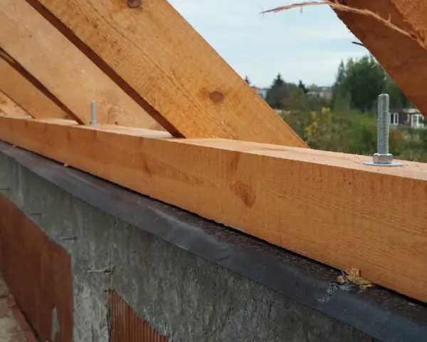Drewniana murłata z widocznymi wymiarami, przykręcona do betonowej ściany. W tle drzewa i domy.