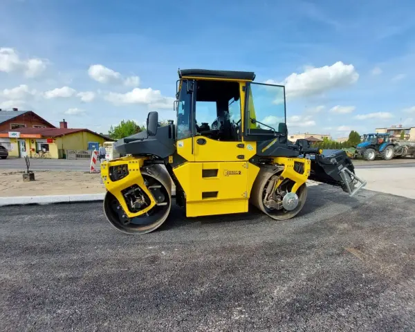Żółty walec drogowy ugniata świeży asfalt. W tle widać budynki i niebieski traktor.