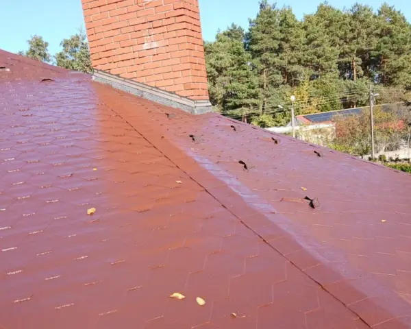 Czerwony dach pokryty gontem bitumicznym z widocznym kominem i drzewami w tle.