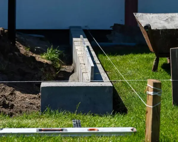 Budowa fundamentu pod ogrodzenie. Betonowe bloczki tworzą solidną podstawę, a sznur wyznacza linię. W tle taczka i zielona trawa.