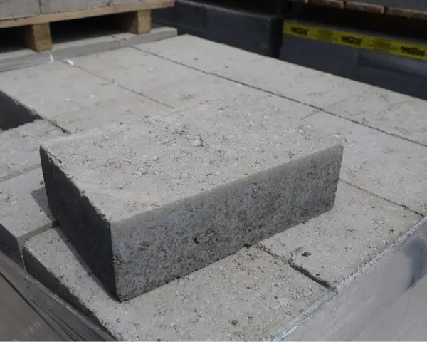 Stos szarych bloczków betonowych, każdy z widocznymi nierównościami i kruszywem. Wymiary bloczków betonowych są standardowe dla budowy.