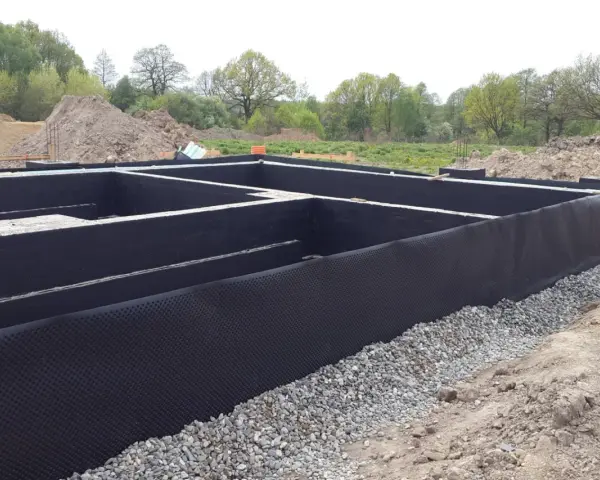 Fundament domu w trakcie budowy, zabezpieczony czarną membraną i otoczony żwirem.