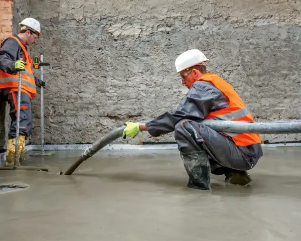 Dwóch budowlańców w kamizelkach odblaskowych wylewa pianobeton. Jeden kieruje wąż, drugi kontroluje poziom.